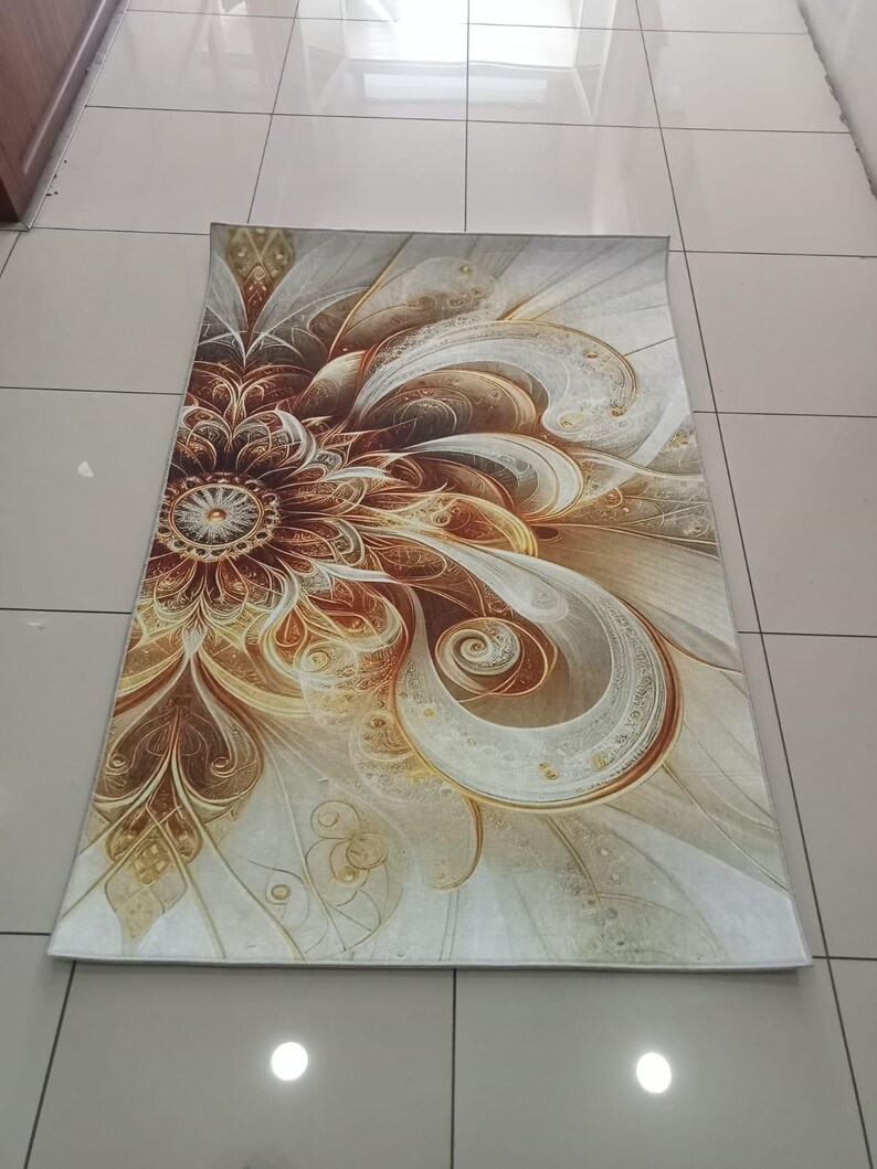 May include: Rectangular rug with a floral design in shades of brown, gold, and cream. The pattern features a central flower with swirling petals and decorative details. The rug is placed on a tiled floor.