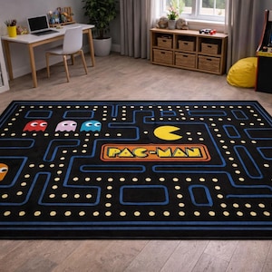 Puede incluir: Alfombra rectangular negra de Pac-Man con un diseño de laberinto. La alfombra presenta el logotipo y los personajes de Pac-Man en amarillo, azul, rojo y rosa. La alfombra está colocada sobre un suelo de madera.