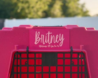 Puede incluir: Un transportín de plástico para mascotas de color rosa brillante con un letrero rectangular que muestra el texto Britney y Afterhours All Eyes On Me en una fuente cursiva rosa claro. Tiene una puerta de rejilla negra y un asa negra.