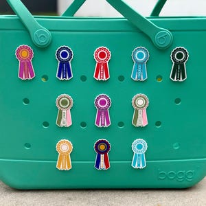 May include: A teal rubber tote bag with the word bogg embossed on the bottom right corner. The bag features twelve circular ventilation holes arranged in a grid, with eleven colorful award ribbon pins attached to the front surface of the bag.