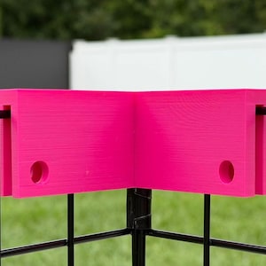 May include: A bright pink L-shaped corner connector piece designed to fit over a black wire grid fence. The connector features two rectangular panels joined at a ninety-degree angle, each with a circular hole near the bottom edge for secure attachment.