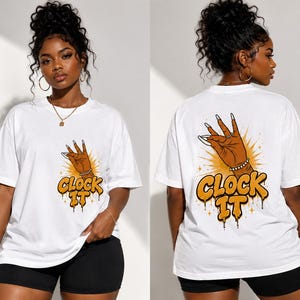 May include: A white short-sleeved t-shirt featuring a graphic print of a hand making a gesture with long nails, accompanied by the text "CLOCK IT" in a bold, dripping orange font with yellow highlights and small star accents on the front and back.