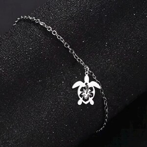 Könnte beinhalten: Ein silberfarbenes Metallkettenarmband mit einem einzelnen Anhänger in Form einer Meeresschildkröte. Der Schildkrötenanhänger hat ein ausgeschnittenes Design, das ein Hibiskusblütenmuster in seinem Panzer darstellt und ein detailliertes Aussehen erzeugt.
