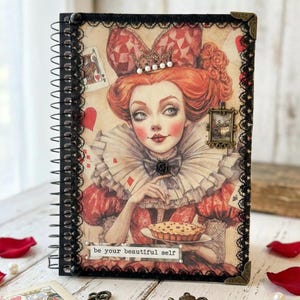 Puede incluir: Un diario con espiral que presenta una ilustración detallada de una mujer con un vestido rojo y una corona, sosteniendo un pastel. La portada está enmarcada con encaje negro y protectores de esquina. El texto "be your beautiful self" está impreso.