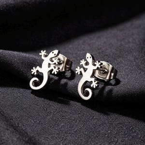 Könnte beinhalten: Ein Paar silberfarbene Ohrstecker in Form kleiner Eidechsen. Jeder Ohrstecker zeigt die flache, metallische Silhouette eines Geckos mit geringeltem Schwanz und detaillierten Füßen, befestigt mit einem standardmäßigen Metallstecker.