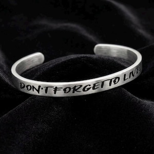 Könnte beinhalten: Ein silbernes Armreif-Armband mit dem eingravierten Text "DON'T FORGET TO LIVE". Das offene Armband ist aus gebürstetem Metall gefertigt und liegt auf einer schwarzen Samtoberfläche. Das Armband ist ein schlichtes, aber aussagekräftiges Schmuckstück.