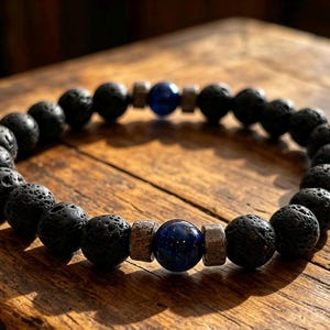 Può includere: Un braccialetto di perline composto da perle rotonde e porose in pietra lavica nera, alternate a due perle sferiche lisce di colore blu intenso. Ogni perla blu è affiancata da due piccoli distanziatori metallici cilindrici con una finitura testurizzata.