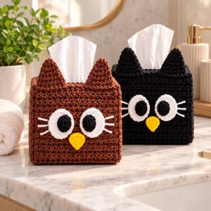 Puede incluir: Dos fundas cuadradas para cajas de pañuelos hechas de hilo de ganchillo marrón y negro. Cada funda presenta orejas puntiagudas tipo gato, grandes ojos circulares blancos y negros y una pequeña nariz triangular amarilla, con pañuelos blancos.