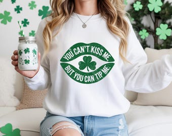 May include: White sweatshirt with a green graphic of lips and a shamrock, with the text "YOU CAN'T KISS ME, BUT YOU CAN TIP ME." A mason jar with a green shamrock design is held in the left hand. The background has green shamrock decorations.