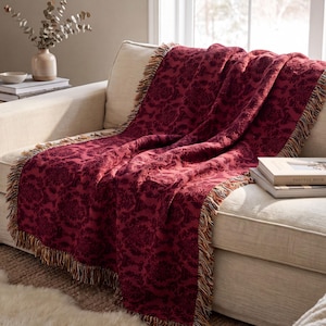 May include: A deep red woven throw blanket featuring a repeating damask pattern and multi-coloured fringe along the edges. The textile has a textured appearance with a rich, dark crimson hue and visible threads in shades of gold and brown at the borders.
