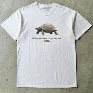 May include: White t-shirt with a tortoise illustration and the text "slow motion over no motion". The National Geographic logo is below the text. The t-shirt is made of a soft material and is suitable for casual wear.