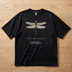 Può includere: T-shirt nera con un'illustrazione dettagliata di una libellula con ali beige e corpo verde. Il testo "why am i so fly" è sotto la libellula, con il logo National Geographic sotto.