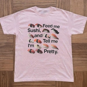 以下が含まれることがあります： 寿司のグラフィックと「Feed me Sushi, and Tell me I'm Pretty」のテキストが入った薄いピンク色のTシャツ。グラフィックには様々な寿司ロールが含まれています。柔らかい素材で作られたシャツです。
