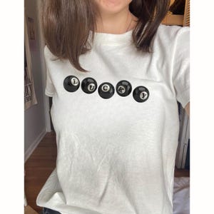 Può includere: T-shirt bianca con la scritta "LUCKY" realizzata con palle da biliardo nere. Le palle sono disposte orizzontalmente. La maglietta è realizzata in un materiale morbido e confortevole.