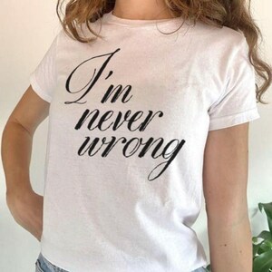 May include: A white short-sleeved t-shirt featuring the black cursive text I'm never wrong printed on the front. The garment has a crew neckline and a standard fit, showcasing the bold lettering clearly against the plain white fabric background.