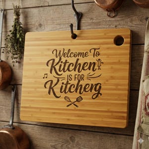 May include: A rectangular bamboo cutting board with the text "Welcome To Kitchen Is For Kitcheng" engraved in a decorative font. The board has a hole in the upper right corner and is hanging on a wooden wall.
