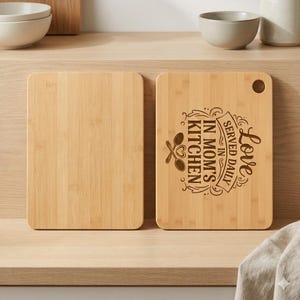 May include: Two rectangular bamboo cutting boards. One is blank, the other features the text "Love Served Daily in Mom's Kitchen" with spoon and heart graphics. Both have rounded corners and a hole for hanging.