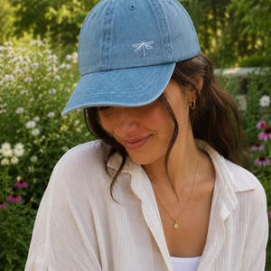 May include: A light blue denim baseball cap featuring a small, white embroidered dragonfly design on the front panel. The cap has a curved brim, a structured crown with ventilation eyelets, and a classic six-panel construction with a button top.