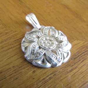 Puede incluir: Un colgante de plata en forma de flor con detalles intrincados. La flor tiene un c&iacute;rculo central con un patr&oacute;n texturizado y est&aacute; rodeado de p&eacute;talos con un borde de cuentas.
