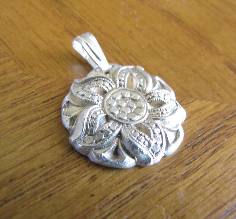Puede incluir: Un colgante de plata en forma de flor con detalles intrincados. La flor tiene un c&iacute;rculo central con un patr&oacute;n texturizado y est&aacute; rodeado de p&eacute;talos con un borde de cuentas.