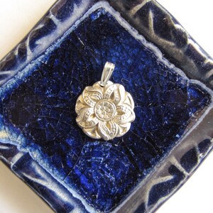 Puede incluir: Un colgante de plata en forma de flor con un dise&ntilde;o detallado, descansando sobre una baldosa de cer&aacute;mica azul y blanca.