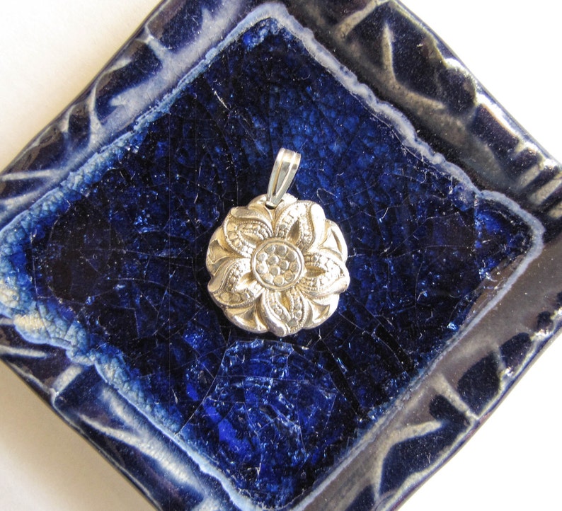 Puede incluir: Un colgante de plata en forma de flor con un dise&ntilde;o detallado, descansando sobre una baldosa de cer&aacute;mica azul y blanca.