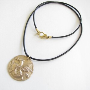 K&ouml;nnte beinhalten: Eine goldfarbene Halskette mit einem detaillierten Drachen-Anh&auml;nger. Die Kette ist aus schwarzem Lederband mit einem goldfarbenen Verschluss.
