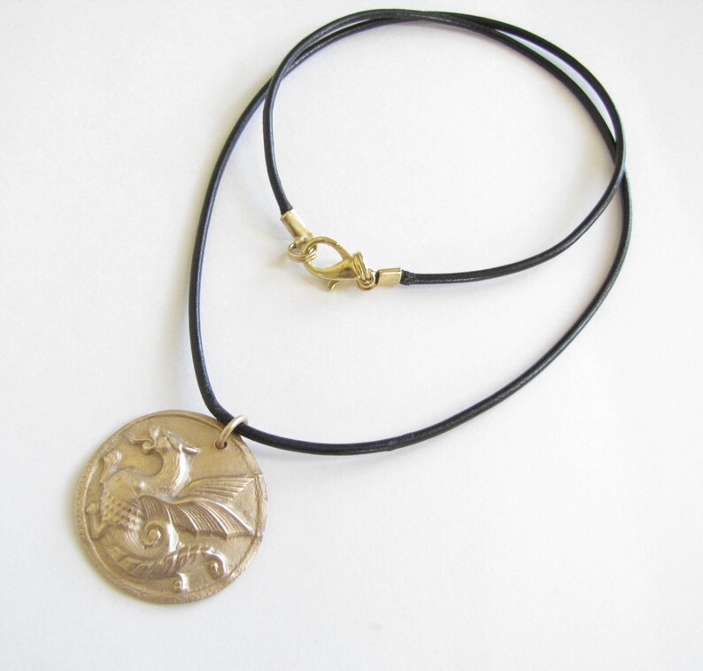 K&ouml;nnte beinhalten: Eine goldfarbene Halskette mit einem detaillierten Drachen-Anh&auml;nger. Die Kette ist aus schwarzem Lederband mit einem goldfarbenen Verschluss.