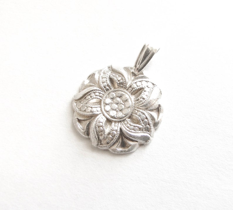 Puede incluir: Un colgante de plata en forma de flor con un dise&ntilde;o detallado. El colgante tiene una forma circular con un dise&ntilde;o de flor de cinco puntas en el centro.
