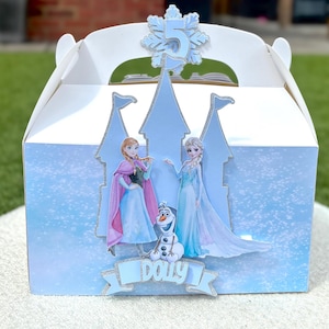 Puede incluir: Una caja de cart&oacute;n blanca decorada con la silueta de un castillo azul claro, con recortes de Anna, Elsa y Olaf. Un adorno de copo de nieve brillante muestra el n&uacute;mero 5, y una cinta azul en la parte inferior presenta el nombre Dolly en blanco.