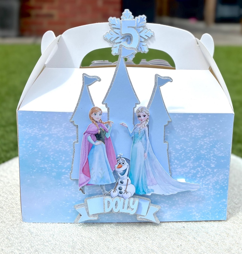 Puede incluir: Una caja de cart&oacute;n blanca decorada con la silueta de un castillo azul claro, con recortes de Anna, Elsa y Olaf. Un adorno de copo de nieve brillante muestra el n&uacute;mero 5, y una cinta azul en la parte inferior presenta el nombre Dolly en blanco.