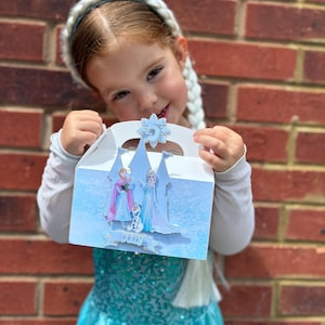 Puede incluir: Una caja de cart&oacute;n blanca con asa, decorada con un dise&ntilde;o invernal en azul claro que muestra la silueta de un castillo, dos personajes femeninos, un mu&ntilde;eco de nieve y un copo de nieve con el n&uacute;mero 5. El nombre Dolly aparece en una cinta.
