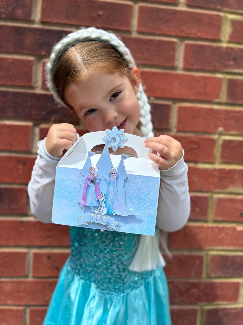 Puede incluir: Una caja de cart&oacute;n blanca con asa, decorada con un dise&ntilde;o invernal en azul claro que muestra la silueta de un castillo, dos personajes femeninos, un mu&ntilde;eco de nieve y un copo de nieve con el n&uacute;mero 5. El nombre Dolly aparece en una cinta.