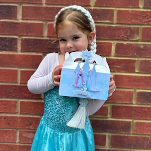 Puede incluir: Una caja de regalo de cart&oacute;n blanco con un patr&oacute;n azul helado y un recorte de un castillo con los personajes Anna, Elsa y Olaf. La caja est&aacute; rematada con un emblema de copo de nieve azul e incluye una borla blanca decorativa colgando.