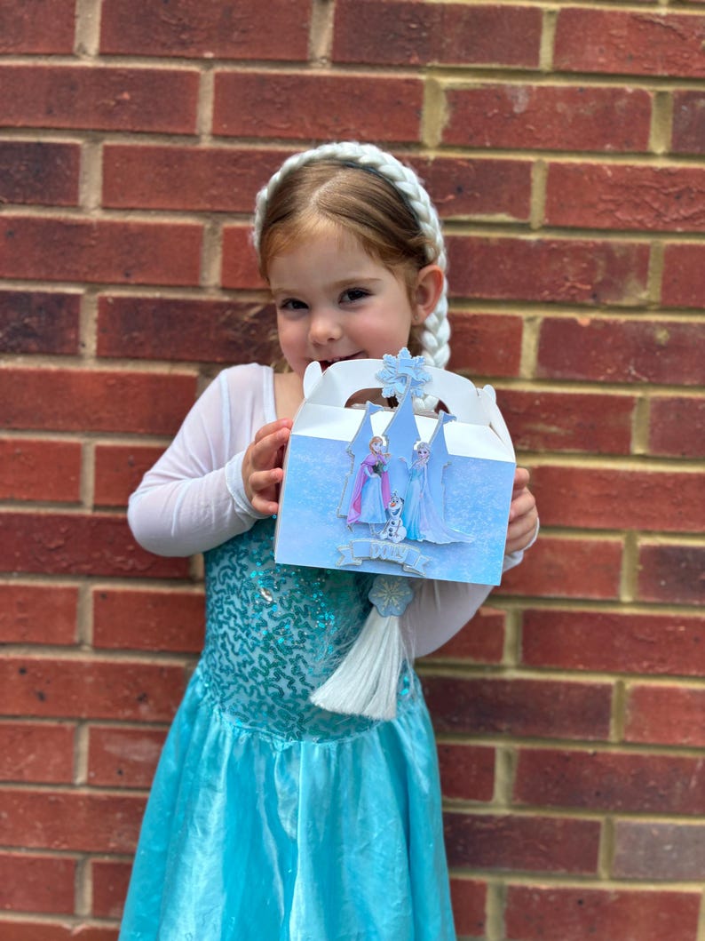 Puede incluir: Una caja de regalo de cart&oacute;n blanco con un patr&oacute;n azul helado y un recorte de un castillo con los personajes Anna, Elsa y Olaf. La caja est&aacute; rematada con un emblema de copo de nieve azul e incluye una borla blanca decorativa colgando.