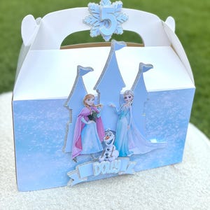 Puede incluir: Una caja de cart&oacute;n blanca con un recorte de castillo azul claro con bordes brillantes. La caja muestra im&aacute;genes de dos personajes animados y un mu&ntilde;eco de nieve, con el nombre Dolly impreso en una pancarta azul y un n&uacute;mero 5 en un copo de nieve.