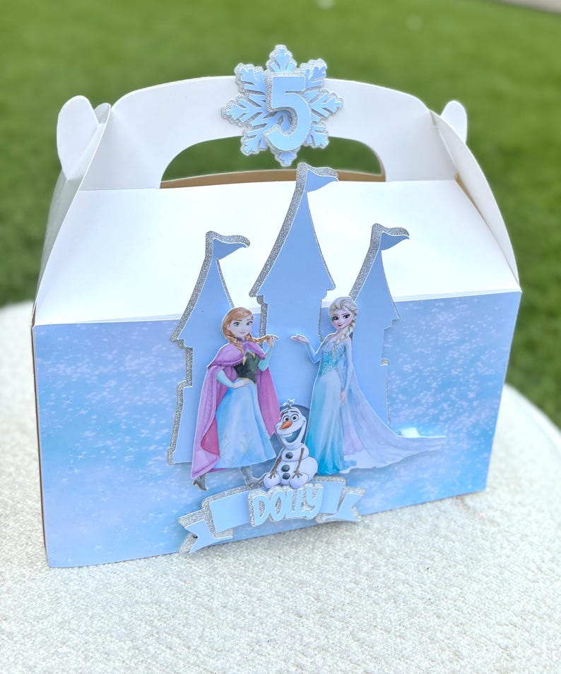 Puede incluir: Una caja de cart&oacute;n blanca con un recorte de castillo azul claro con bordes brillantes. La caja muestra im&aacute;genes de dos personajes animados y un mu&ntilde;eco de nieve, con el nombre Dolly impreso en una pancarta azul y un n&uacute;mero 5 en un copo de nieve.