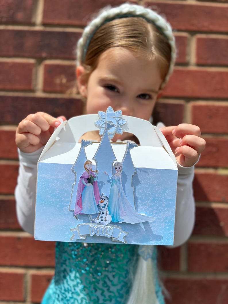 Puede incluir: Una caja de cart&oacute;n blanca con una silueta de castillo azul claro, im&aacute;genes de dos personajes femeninos y un mu&ntilde;eco de nieve, un recorte de copo de nieve con el n&uacute;mero 5 y una pancarta con el nombre Dolly impreso en letras azules.