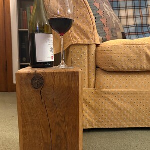 May include: A bottle of wine and a glass of red wine are on a square wooden block. The block has a natural wood grain and a knot. The background includes a couch with a patterned cushion and a bookshelf. The block is approximately 30 cm in height.