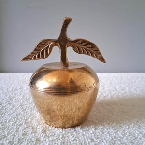 May include: A golden-colored apple-shaped trinket box with a stem and leaf design. The lid of the box is adorned with a detailed stem and two leaves. The box has a smooth, reflective surface.