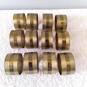 May include: A set of twelve brass napkin rings. Each ring is cylindrical with vertical and horizontal striped detailing. The rings are arranged on a white surface, suggesting a dining or table setting theme.