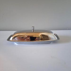 May include: A rectangular butter dish made of polished stainless steel with a smooth, reflective surface. The dish features a removable lid with a small, centered knob handle and a base with slightly raised edges for a secure fit.