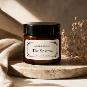 以下が含まれることがあります： 「Elleback Skincare The Sparrow Creamy Serum」とラベルされた、黒い蓋が付いた茶色のガラス瓶。瓶は、ニュートラルカラーの布と背景にドライフラワーのある石の表面に置かれています。ラベルには装飾的な花の要素があります。