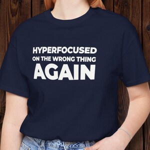 May include: Navy blue t-shirt with the white text "HYPERFOCUSED ON THE WRONG THING AGAIN." The shirt is a crew neck style and appears to be made of a soft material. The shirt is being worn by a person.