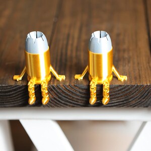 May include: Two small, gold-colored, bullet-shaped figurines with silver-toned tips. Each figurine features articulated arms and legs, allowing them to sit upright with their feet dangling over the edge of a flat, dark brown wooden surface.