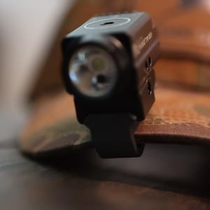 May include: A black tactical flashlight attached to a camouflage hat. The flashlight has a bright LED light and is mounted with a black clip. The hat has a brown and green camouflage pattern.
