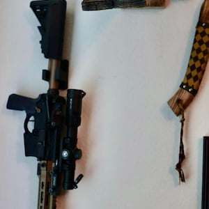 May include: An AR-15 style rifle with a scope and tactical accessories is mounted on a white wall. Above it is a deer skull mounted on a wooden plaque, and a decorative item.