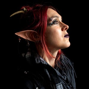 May include: A woman with red hair and elf ears wearing a black jacket and a scarf. She has dark eye makeup and a piercing above her eyebrow.