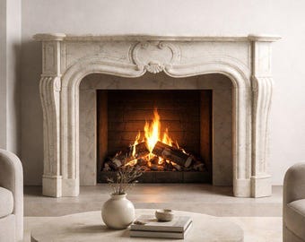 May include: A light beige stone fireplace mantel with carved detailing and a central shell motif, framing a brick-lined hearth with a burning wood fire. A round stone coffee table sits in the foreground with a small vase and stacked books.
