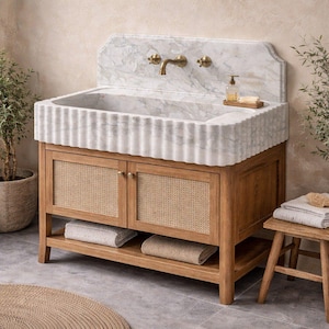 Può includere: Un lavabo in marmo con bordo scanalato è posto su un mobile in legno con pannelli porta in canna intrecciata. Il lavabo ha rubinetti in ottone. Un sgabello in legno, asciugamani, una piccola pianta e un tappeto sono anche nell'immagine.